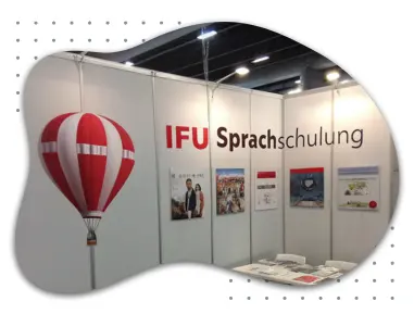 Ausstellungsstand der IFU Sprachschulung mit rotem Heißluftballon, Informationsmaterialien und Plakaten. Der Stand zeigt die IFU-Sprachschule und deren Angebote auf einer Messe.