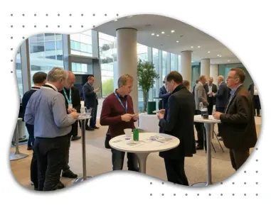 Professionelle Konferenz mit Teilnehmern, die in Gespräche vertieft sind. Gruppen von Personen stehen an hohen Tischen, tauschen Visitenkarten aus und diskutieren in einem modernen, hellen Konferenzraum mit großen Fenstern.