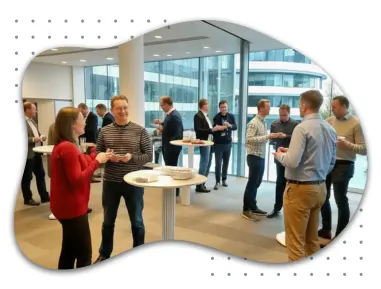 Participants at a professional conference engaging in discussions. Several groups of people stand around high tables, chatting and exchanging business cards in a modern conference space with large windows and a bright atmosphere.